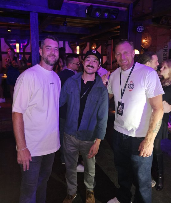 Three men smiling and posing for a photo at what appears to be a social event or party in a venue with purple lighting.