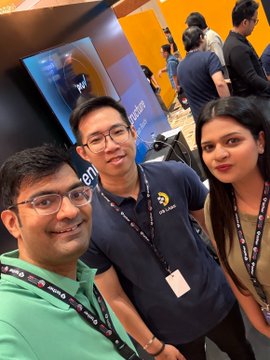 A selfie of three people at what appears to be a tech event or office, including individuals wearing glasses and ID lanyards in front of computer monitors.