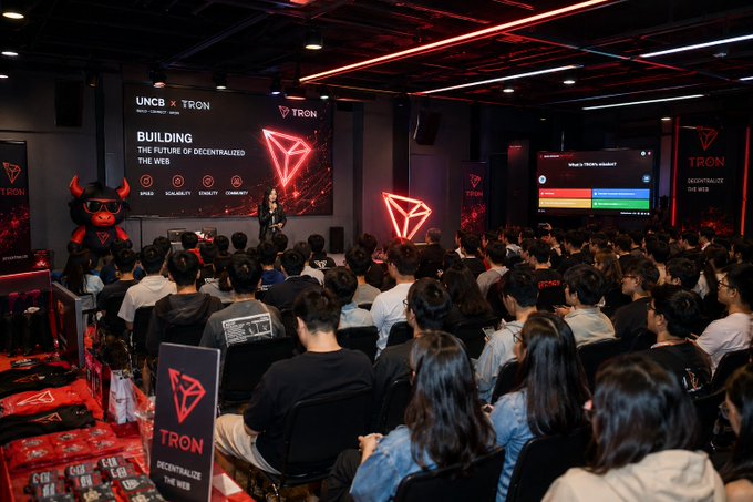 A packed audience attends the UNCB × TRON event, with the stage screen displaying "BUILDING THE FUTURE OF DECENTRALIZED THE WEB" alongside the TRON logo, as the post emphasizes that strong attendance is key to sparking curiosity and turning newcomers into future builders within the ecosystem.