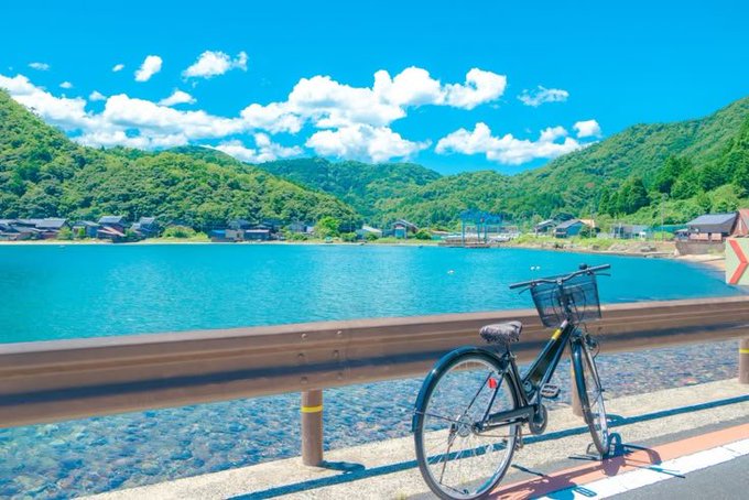 A black bicycle leans against a metal guardrail overlooking a vibrant turquoise bay surrounded by green mountains and a small coastal village under a sunny sky.