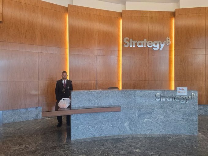 A security guard stands in the wood-paneled lobby of Strategy's headquarters behind a marble reception desk featuring the company's "Strategy" logo, with what appears to be a FedEx package of oranges on the counter. The image humorously illustrates the post's reference to delivering oranges to Michael Saylor's office as a playful jab about Bitcoin (referred to as the "orange coin").