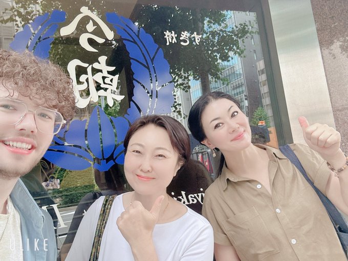 The image shows three people posing for a photo outside of a restaurant with a blue sign that says "Shawn from Orbs' farewell lunch." The group consists of two women and one man, all smiling and enjoying their time together. They are standing in front of a building, which appears to be a restaurant or cafe. One person is holding up their hand for the camera while giving a thumbs-up gesture.