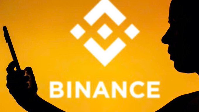 A silhouette of a person holding a smartphone is shown against an orange backdrop featuring the white Binance logo and the text "BINANCE," visually representing the crypto exchange whose research arm reported that stablecoin-settled perpetual contracts tied to traditional financial assets surpassed $1.1 trillion in trading volume.