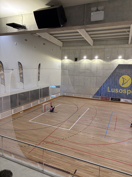 An indoor sports facility with wooden flooring, a goal net, and a partial "Lusosp" logo on the wall, with a person visible near the goal.