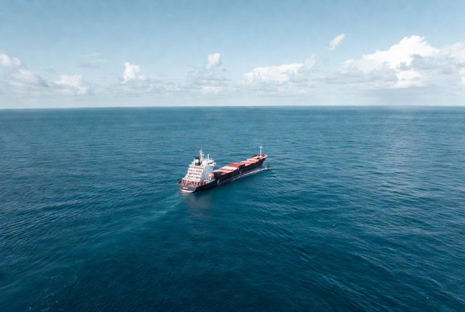 An aerial photograph of a large cargo container ship sailing through deep blue ocean waters under a bright sky with scattered clouds.