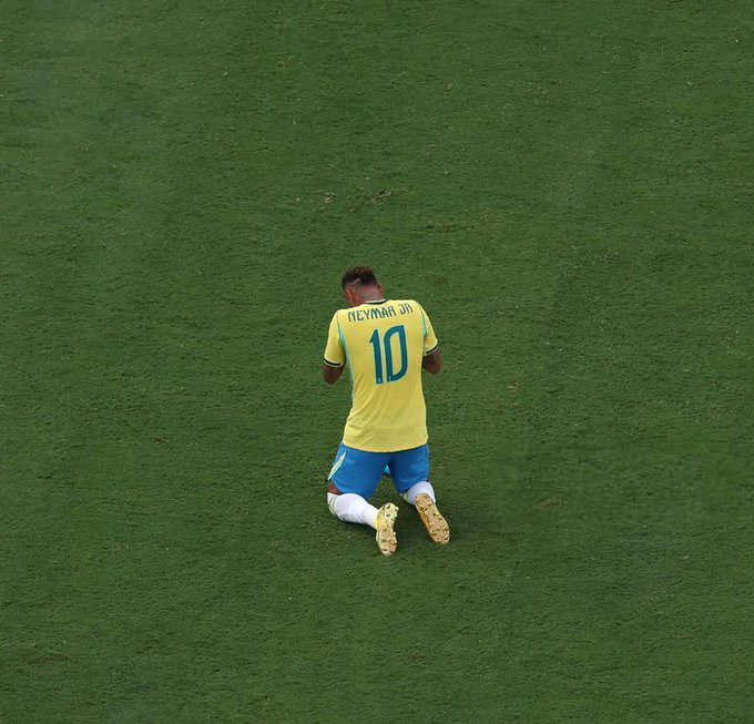 An aerial view of Brazilian soccer player Neymar Jr. (number 10) kneeling alone on a green grass field in his yellow and blue kit, captured from a bird's-eye perspective.