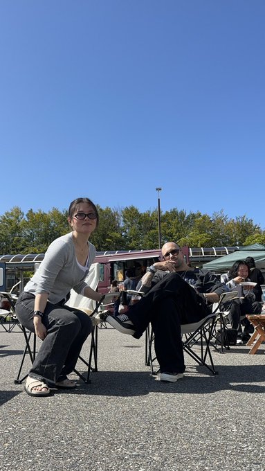 Two women sit in folding camp chairs at an outdoor event on a sunny day, one wearing sunglasses and a gray top, the other in a dark jacket holding a drink, with food trucks visible in the background.
