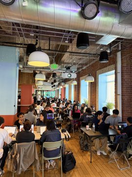 A group of people gather at long tables in a modern, industrial-style event space with exposed brick walls and hanging pendant lights, participating in the VoiceOS hackathon. The setting reflects the collaborative and energetic atmosphere of the coding event where teams competed to win awards, including "best use of Convex."