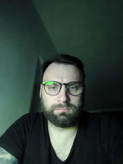 The image features a man wearing glasses with a beard and looking directly at the camera. The background appears to be a dark green color, which contrasts with the man's black t-shirt. There are no texts or other objects present in the image.