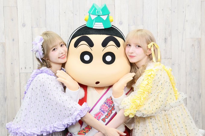 The image features two women posing next to a large, anthropomorphic stuffed animal statue of a character named "Luna" wearing a green hat. The woman on the left has purple hair and is wearing a white dress with purple flowers, while the other woman on the right has blonde hair and is dressed in yellow. They are both smiling for the camera, creating an endearing scene that showcases their friendship or connection to the character "Luna".