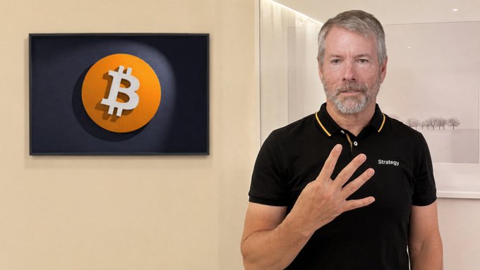 A man with gray hair and beard wearing a black polo shirt with 'Strategy' text stands next to a framed picture of a Bitcoin (₿) logo on a wall, gesturing with his hand.