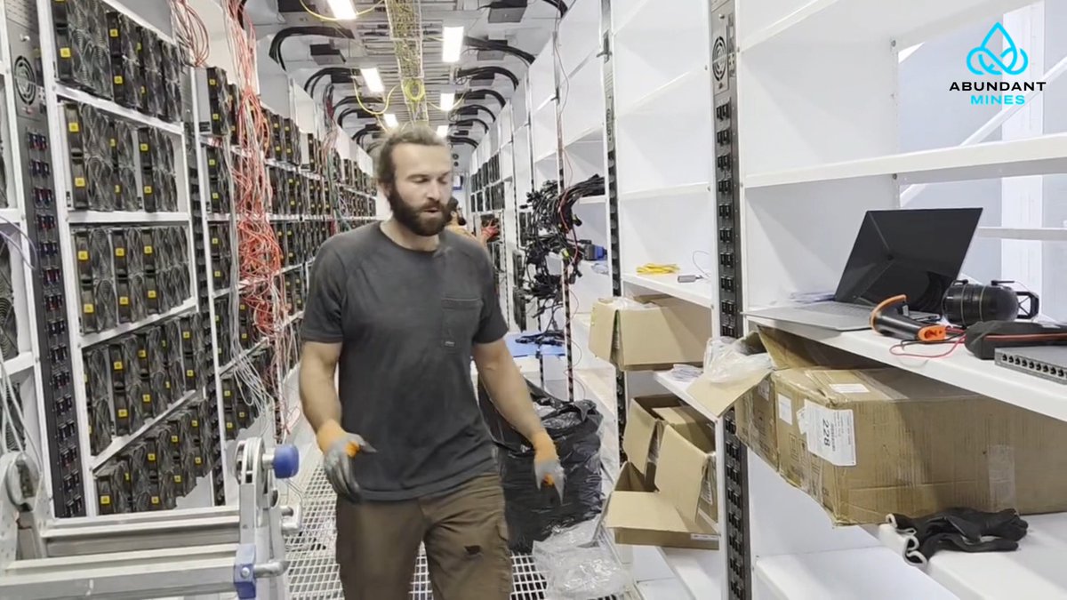A bearded man in a gray t-shirt and work gloves walks down the aisle of a large cryptocurrency mining facility filled with rows of hardware, with 'ABUNDANT MINES' logo in the corner.