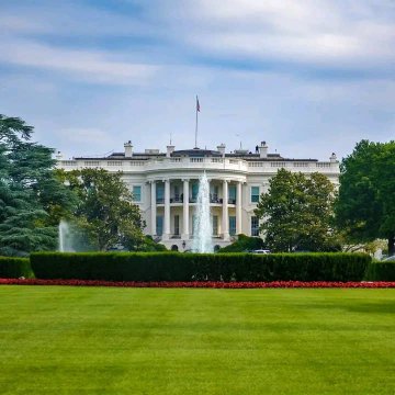 The White House stands under a partly cloudy sky with a manicured lawn and fountain in the foreground.