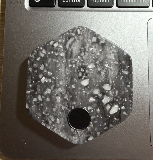 A hexagonal coaster or decorative item with a gray and white terrazzo-like speckled pattern and a black circular button in the center, resting on a silver laptop surface near the keyboard.