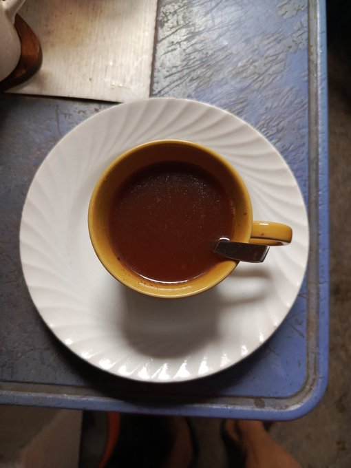 A top-down view of a yellow ceramic cup filled with a dark reddish-brown beverage (likely tea or coffee) on a white ridged saucer, sitting on a worn blue table.