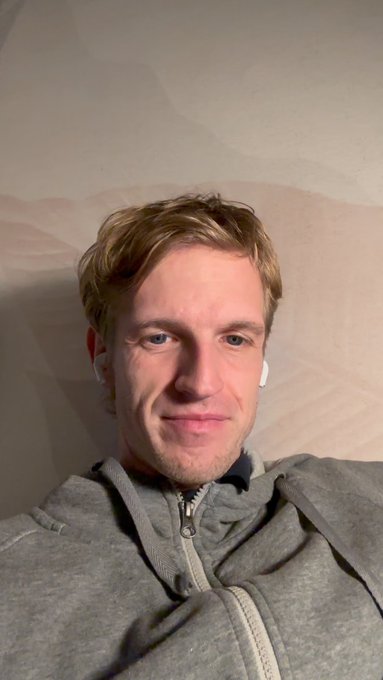A man with blond hair, wearing a gray hoodie and wireless earbuds, takes a relaxed selfie while reclining against a pillow, presumably posting from Vienna. The casual, intimate photo aligns with the post's reflective Sunday message about shared struggles with agentic software engineering, offering a personal glimpse into the speaker's morning routine.