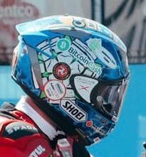 The image features a person wearing a blue helmet with various stickers on it. The helmet has a sticker that says "BITTECHO" and another one that reads "SHOEI". There are also other stickers on the helmet, including one for Bitcoin Cash. The person is standing against a blue background, which could be a wall or a building. The image does not provide any information about the post's content, but it seems to be related to the racing world as there is a reference to "BCH" and "Deano FTW!".
