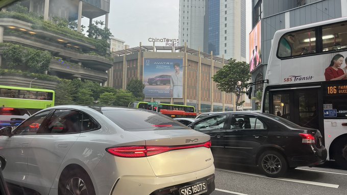 A street-level photograph shows heavy traffic in Singapore, featuring a white BYD sedan with license plate 'SNS1807S', a black car, and an SBS Transit double-decker bus, with the 'ChinaTown Point' building visible in the background alongside an advertisement.