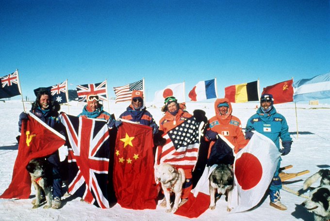 A group of expedition members in winter gear stand in a snowy landscape holding various national flags, including the UK, US, China, Japan, France, Belgium, and USSR, with sled dogs nearby.