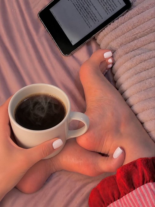 A person holds a steaming white mug of coffee in one hand while using their foot to prop up an e-reader displaying text, all from the cozy perspective of being tucked into pink-striped sheets with a fuzzy blanket nearby. The caption "coffee and toes" aptly captures this cozy morning scene, highlighting the playful pairing of the warm beverage and the pedicured toes visible in the frame.