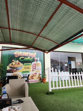 An outdoor covered patio area with a green mesh canopy, white picket fencing, artificial grass, and a banner advertising 'Skazka Cafe' featuring food and drink items.