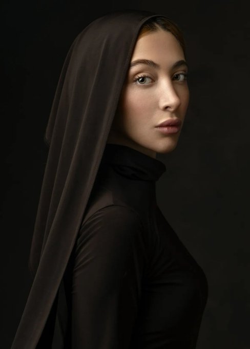 A studio portrait of a woman with light eyes wearing a black long-sleeve turtleneck top and a dark veil draped over her head, set against a black background.