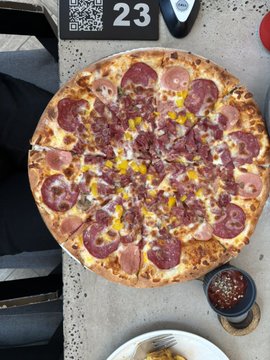 A top-down photograph of a meat-lover's pizza topped with pepperoni, sausage, and corn on a table, with a small cup of sauce beside it and a QR code menu showing the number '23'.