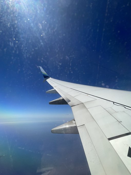 A view from an airplane window showing the wing against a bright blue sky with sunlight streaming through, and clouds visible below.