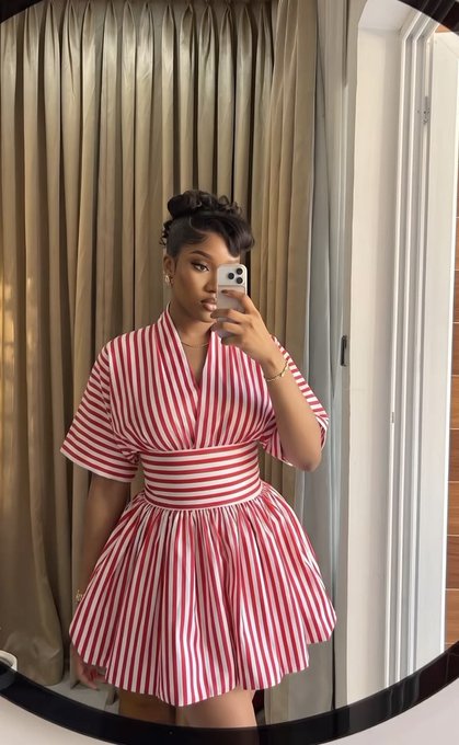 A woman takes a mirror selfie wearing a short, red-and-white striped dress with a matching tie at the neck and her hair styled in a high bun.