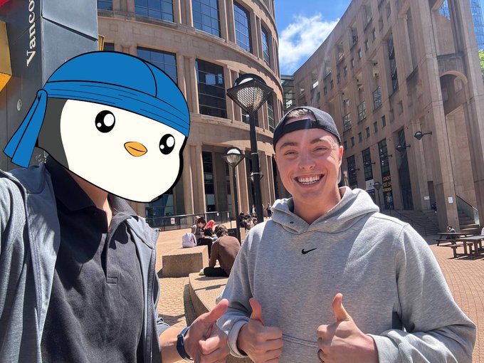 Two people pose outdoors near a building with a Vanco sign; one person's face is covered by a cartoon penguin emoji sticker, while the other smiles and gives a thumbs up.