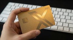 A hand holds a gold-colored credit card with an "X" design on it, in front of a white keyboard. The image illustrates the poster's frustration about being unable to obtain the elusive X (formerly Twitter) branded payment card referenced in their "#XMoney" post.