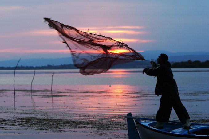 A silhouette of a man standing in a small boat casts a large fishing net against a stunning sunset sky over a calm body of water.