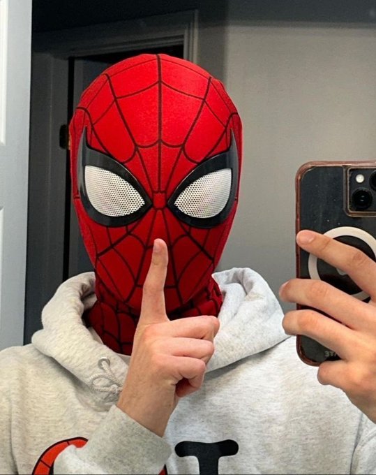 A person wearing a red Spider-Man mask takes a mirror selfie, holding up a peace sign while wearing a gray hoodie.