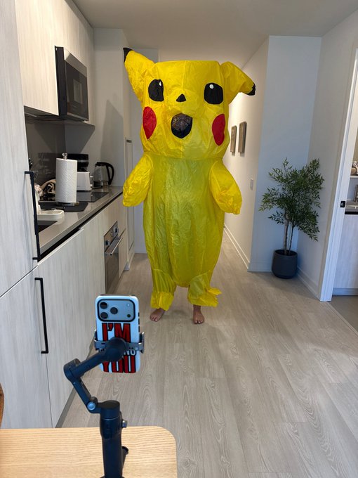 A person in a yellow Pikachu costume stands in a kitchen while a phone on a tripod records them.