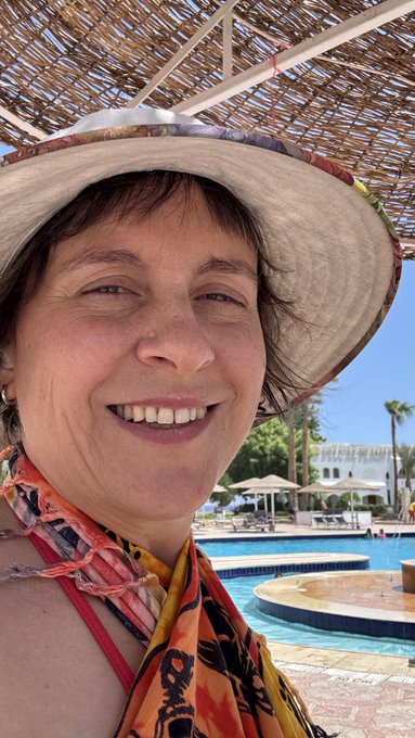 A close-up selfie of a smiling woman wearing a wide-brimmed sun hat and a colorful patterned coverup, taken outdoors at a resort pool area.