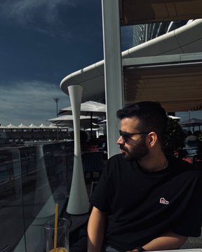 A photo of a bearded man wearing sunglasses and a black t-shirt sitting at an outdoor cafe table under a large umbrella on a sunny day.