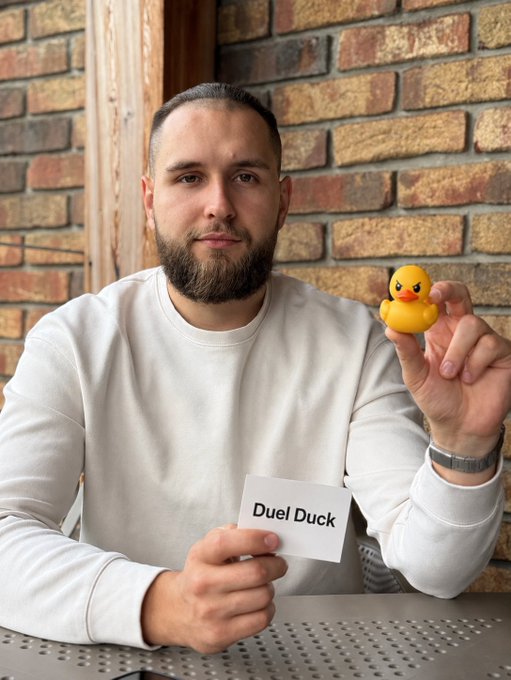A bearded man in a white sweatshirt holds a small yellow rubber duck in one hand and a white card reading 'Duel Duck' in the other, sitting in front of a brick wall.