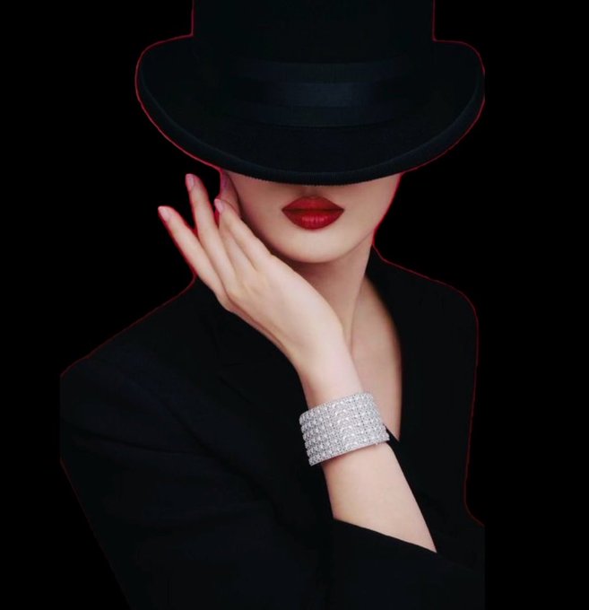 A stylish editorial photo of a woman wearing a black fedora and black blazer, her hand delicately resting against her cheek, showcasing a wide, sparkling diamond bracelet and bold red lipstick against a black background.