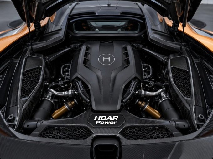 The image shows a close-up view of an engine compartment of a sports car with a black exterior. The engine appears to be well-maintained, and there are two visible exhaust pipes coming out from under the hood. The car is parked on a street or parking lot, as indicated by the presence of a few people in the background who seem to be observing it closely.