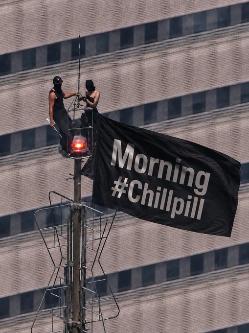 Two people in masks on top of a street pole deploying a black banner reading 'Morning #Chillpill' against a tall office building.