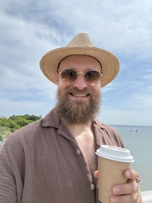 A bearded man wearing a straw sun hat, sunglasses, and an open brown linen shirt smiles while holding a takeaway coffee cup at a sunny beachside. The image has no visible text, but pairs with the caption "GM to future $30k bottom buyers" to evoke a relaxed, optimistic "good morning" greeting aimed at potential crypto or stock market dip-buyers.