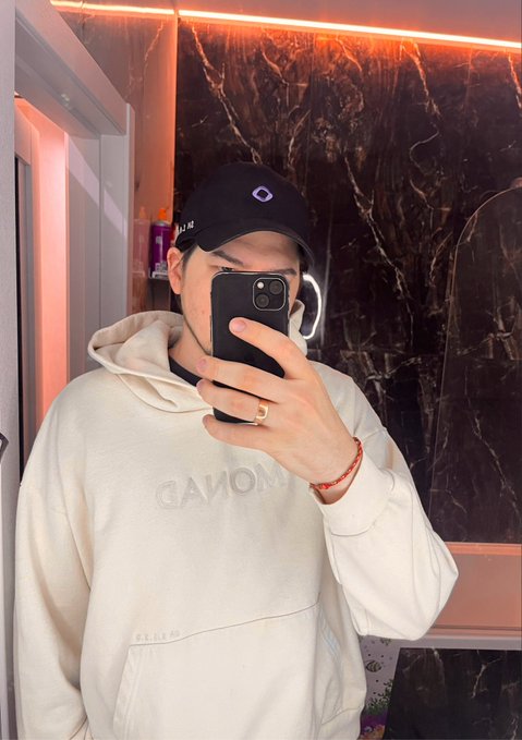 A person takes a mirror selfie wearing a white Canon hoodie and a dark cap, obscuring their face with a phone and wearing a gold ring and red bracelet.