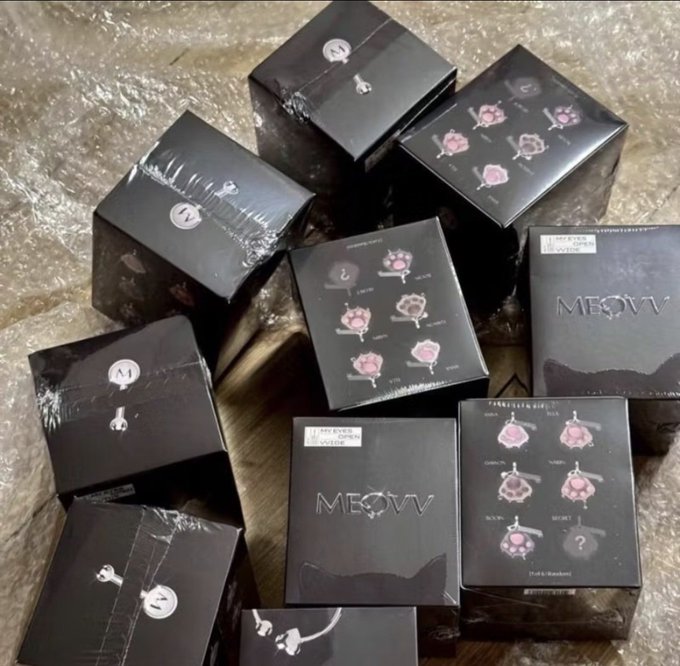 Multiple black MEOVV branded boxes piled together, appearing to be merchandise or album packages with pink floral design elements.
