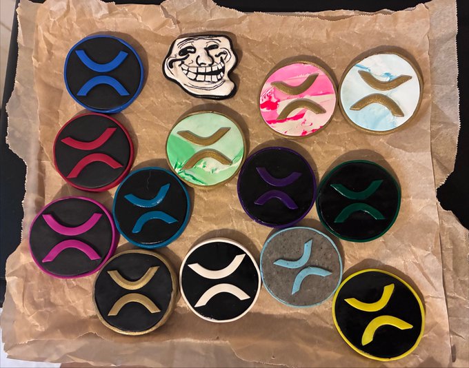 An arrangement of colorful circular cookies or coasters featuring various designs, mostly displaying the XRP (X) cryptocurrency logo, along with one trollface design, resting on brown parchment paper.