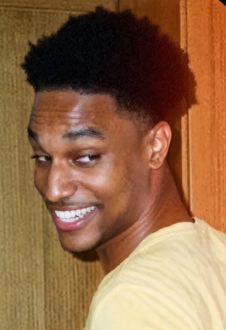 A young man with a high-top fade haircut smiles while looking over his shoulder against a wood-paneled background.