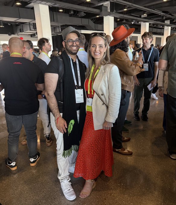 A woman in a white blazer and red dress poses with a man in a black shirt and cap at the Robinhood Summit 2026, both wearing lanyards and smiling in a crowded venue. The hashtags and tagged accounts in the post suggest this gathering of crypto-focused creators and investors.