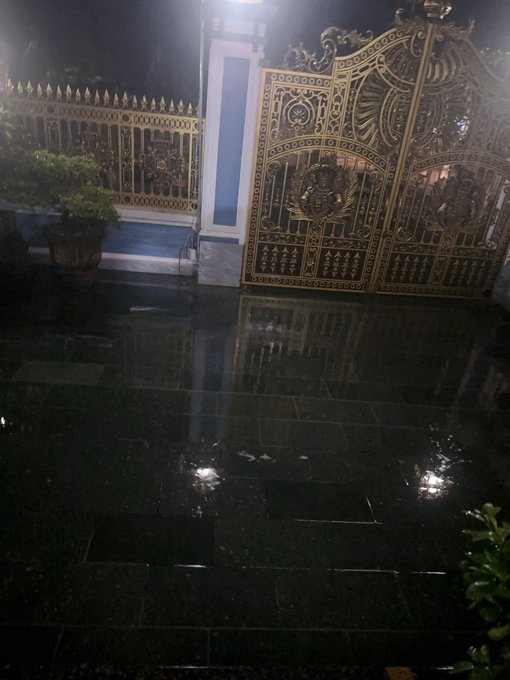 An ornate gold-colored decorative metal gate and fence illuminated at night, reflected on a wet dark tiled surface.