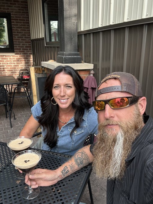 A selfie of a smiling woman with long dark hair and a bearded man wearing sunglasses and a cap, sitting at an outdoor table with two espresso martinis.