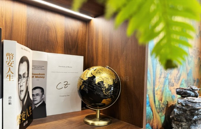 A wooden bookshelf displaying books by Yu 'CZ' Bi, including 'Freedom of Money' (尚安人生) alongside a black and gold globe and decorative plants.