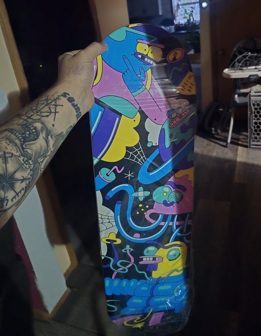 A tattooed hand holds up a skateboard deck featuring a colorful, psychedelic cartoon design with characters and abstract shapes.